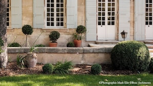 Ein historisches Gebäude mit Bogenfenstern und Fensterläden, umgeben von einem gepflegten Garten mit Topfpflanzen und einer ordentlich geschnittenen Hecke.