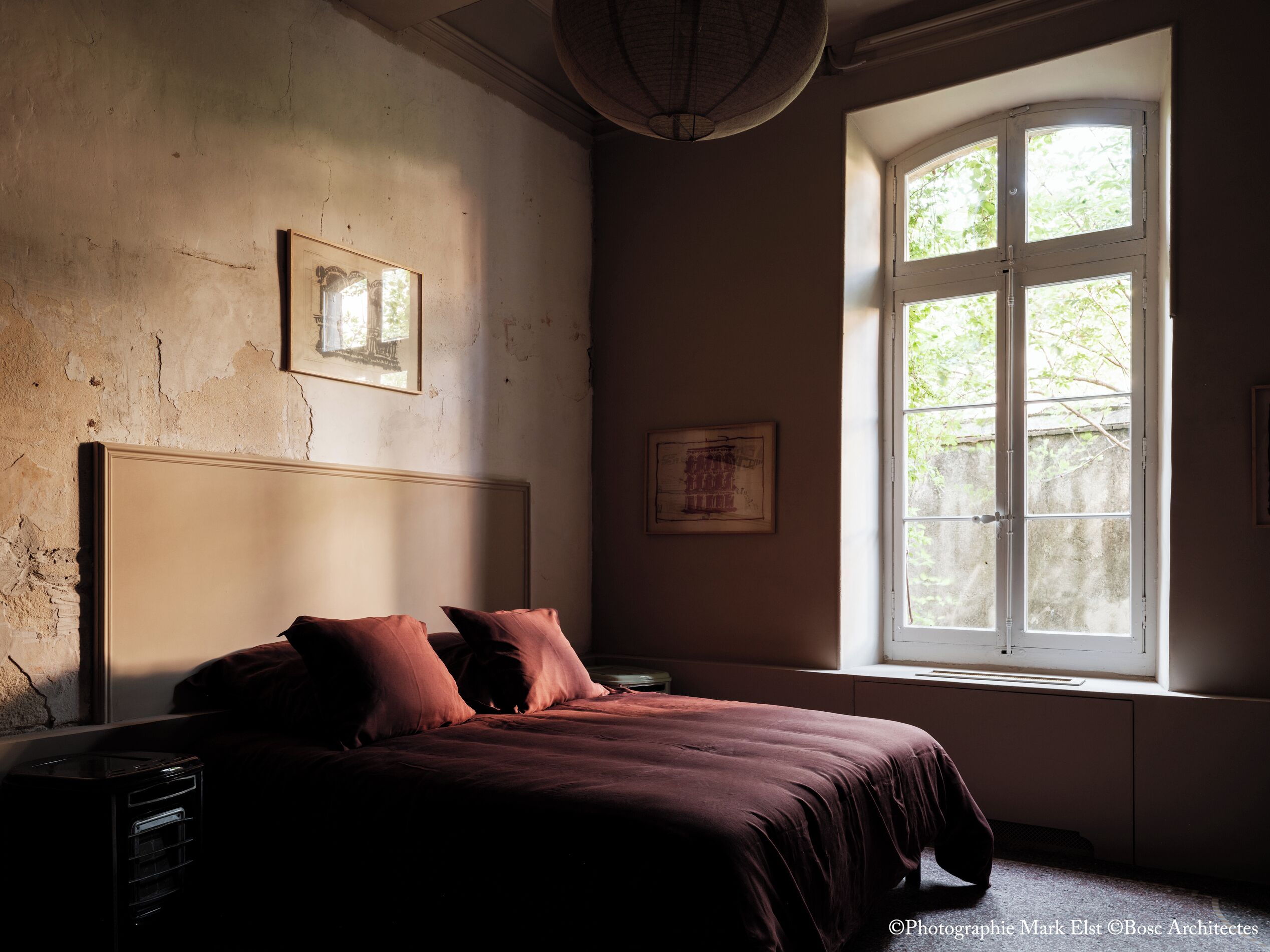Une chambre avec une grande fenêtre, un lit doté d’une tête de lit et deux tableaux encadrés accrochés au mur.