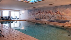 An indoor swimming pool with a mural of mountains and clouds on the wall, lounge chairs along the side, and a skylight above the pool.