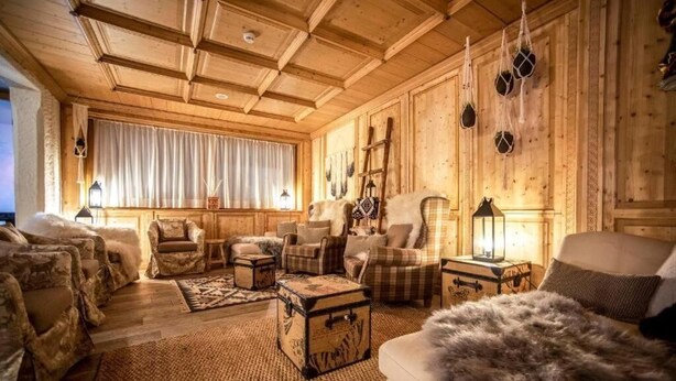A cozy log cabin interior with wooden walls, a large window with curtains, and a variety of seating including sofas and armchairs.