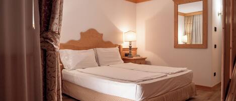 A neatly made bed in a room with wooden flooring, a nightstand with a lamp, and a mirror on the wall.