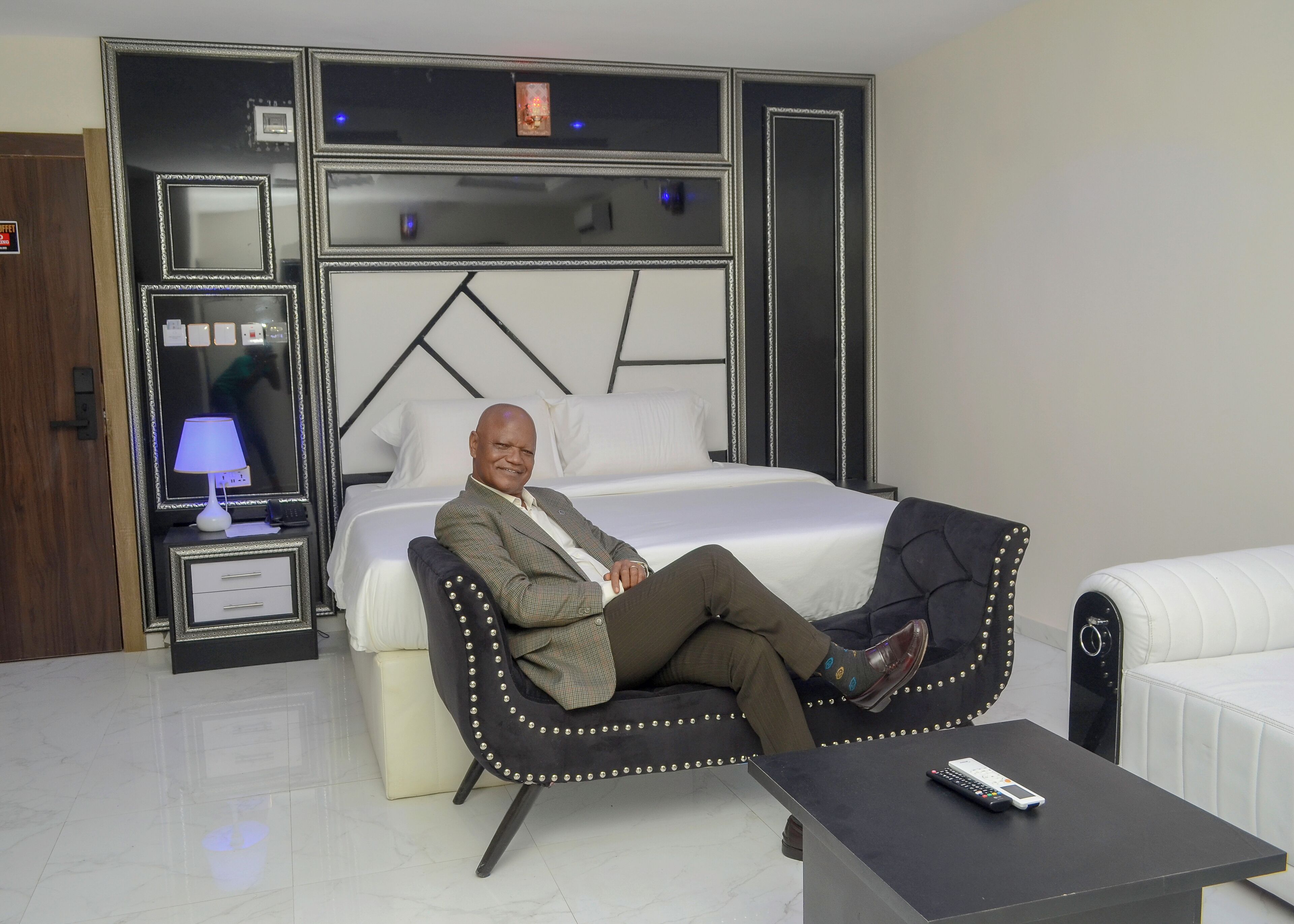 A hotel room with a bed, a bedside table with a lamp, a chair, a small table, and a remote control.