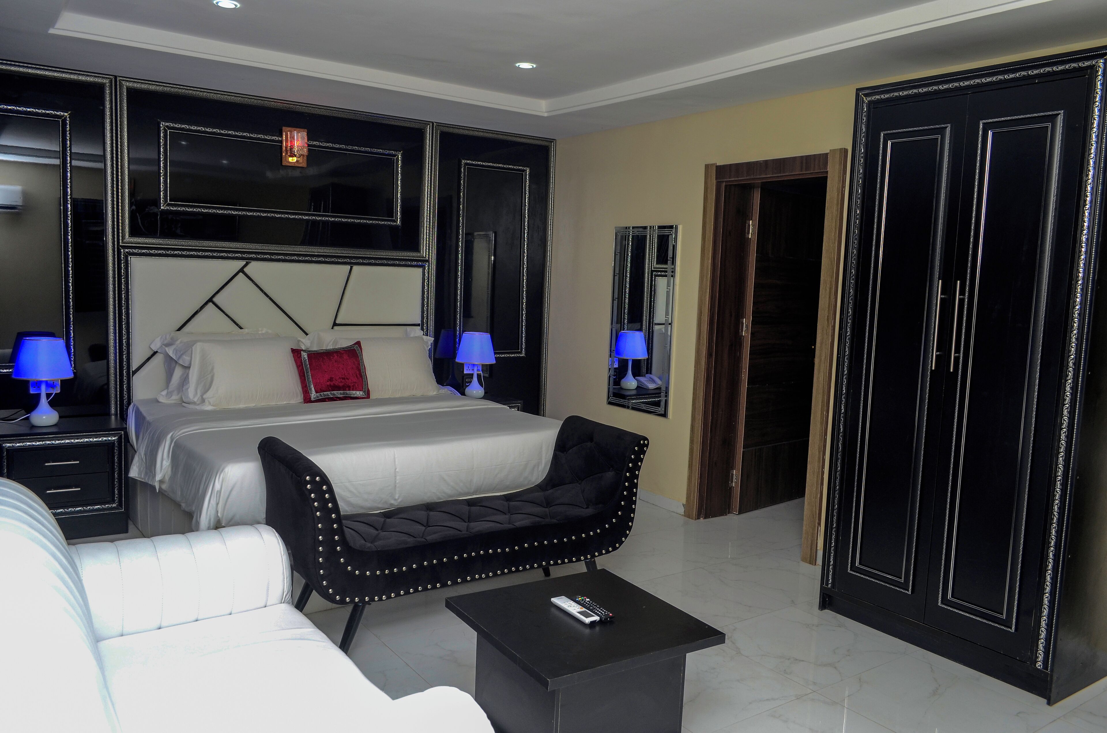 Un dormitorio moderno con una cama grande, mesitas de noche con lámparas, un banco tapizado en negro, una pequeña mesa negra con un control remoto y un armario con puertas espejadas.