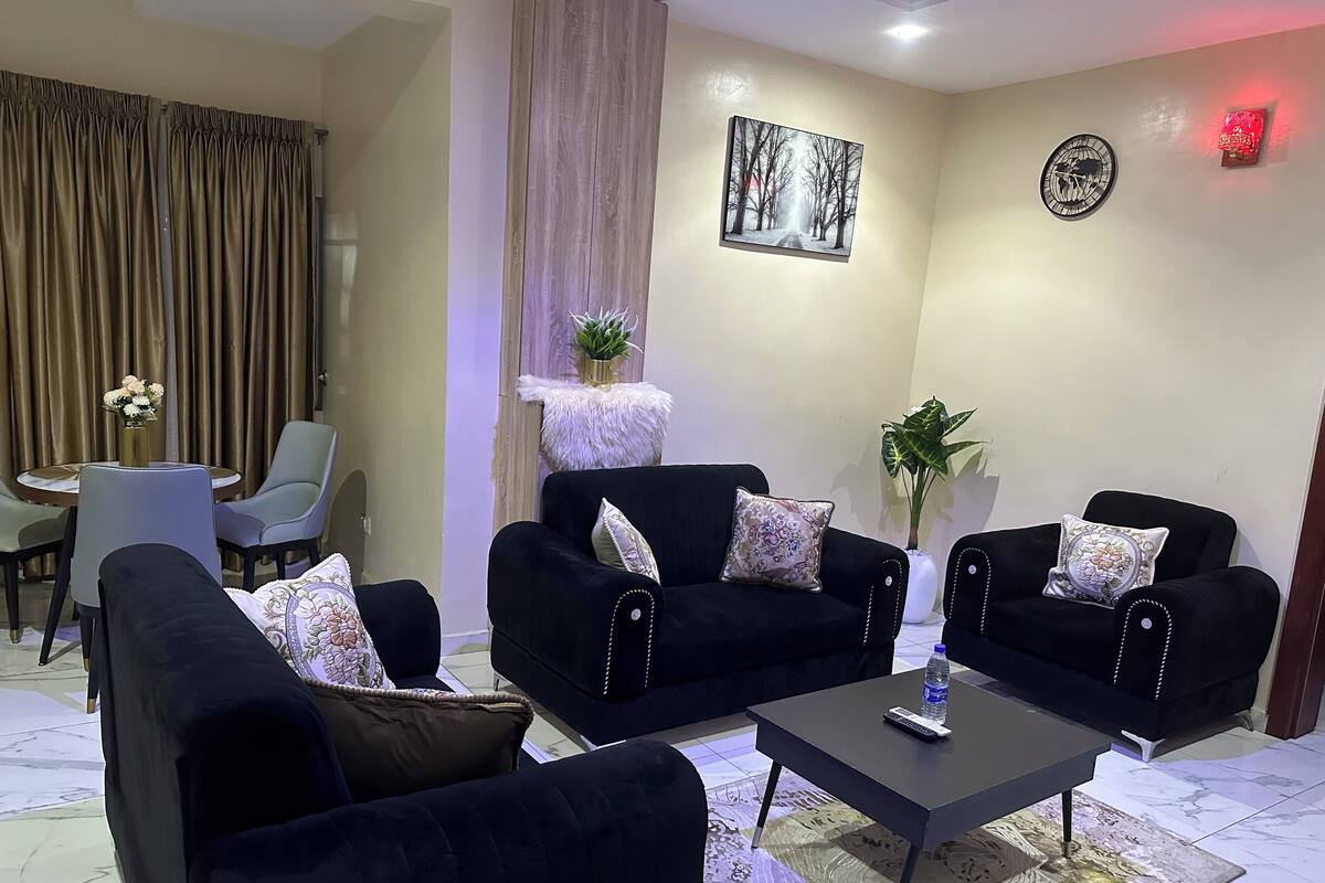 Un salon avec deux canapés noirs, une table basse, une petite table avec des chaises, une plante en pot et un tableau accroché au mur.