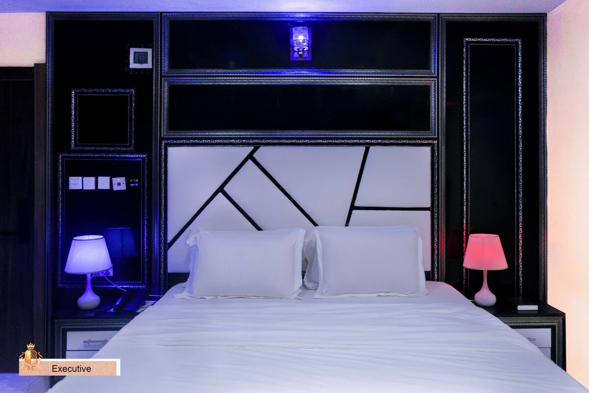 Une chambre d’hôtel avec un grand lit, deux tables de chevet, une petite table, un panneau de commande fixé au mur et un luminaire au plafond.