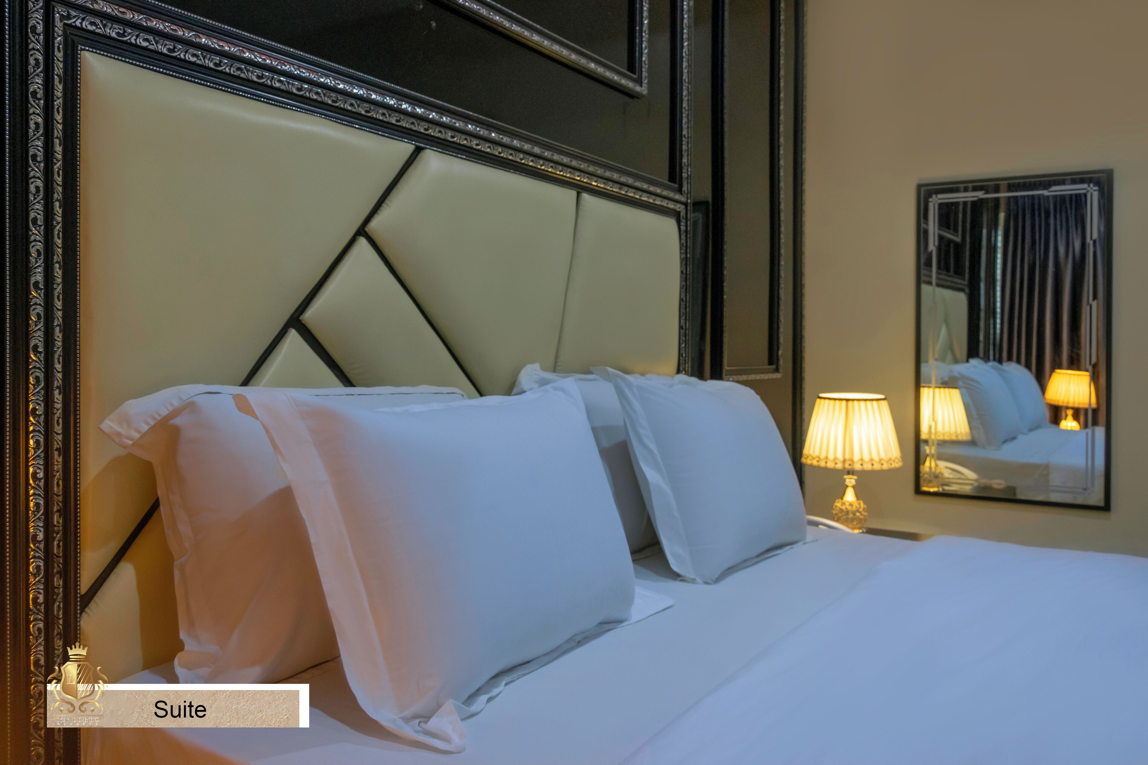 A hotel suite with a bed, pillows, a nightstand with a lamp, and a mirror.