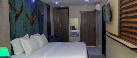 A modern hotel room with a large bed, a bedside table, a mirror, a TV, and a wall-mounted shelf.