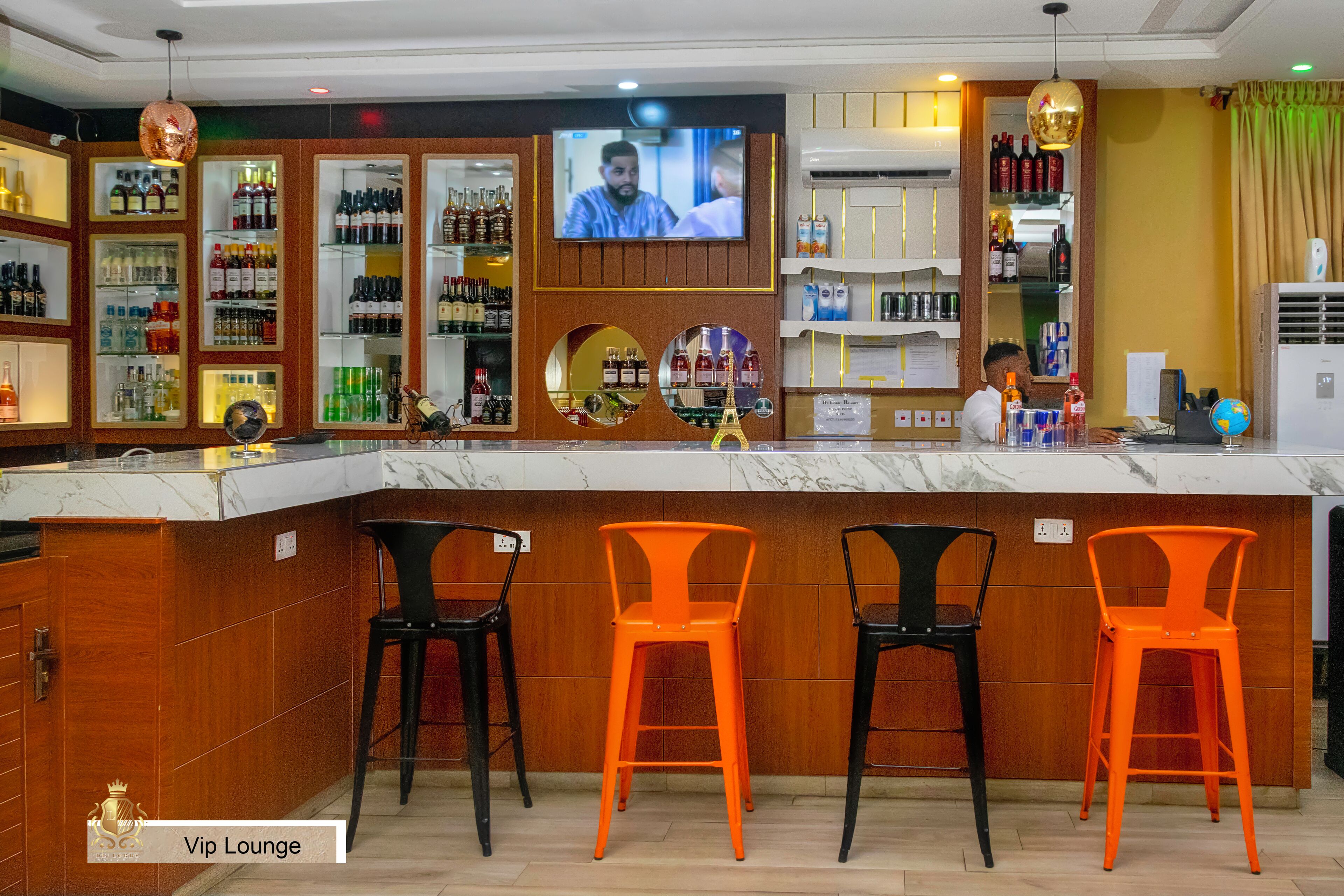 Zona de bar con una pantalla de televisión, estantes con botellas y tres sillas naranjas.