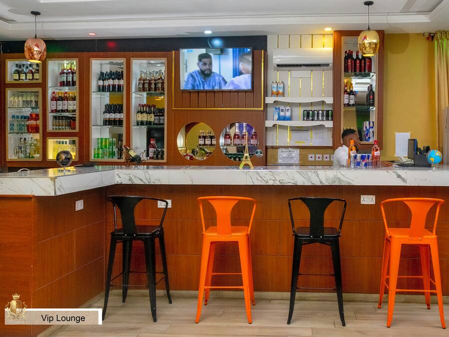 Zona de bar con una pantalla de televisión, estantes con botellas y tres sillas naranjas.