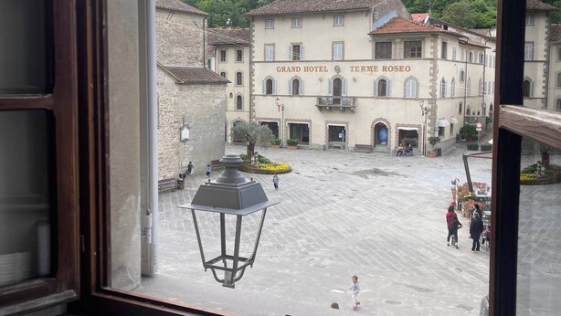 A view from a window overlooking a courtyard with a Grand Hotel Termé Roseo building, stone buildings, and a forested hill. - Tranquil 2-bedroom Apartment in Historic Bagno di Romagna (Bagno di Romagna)