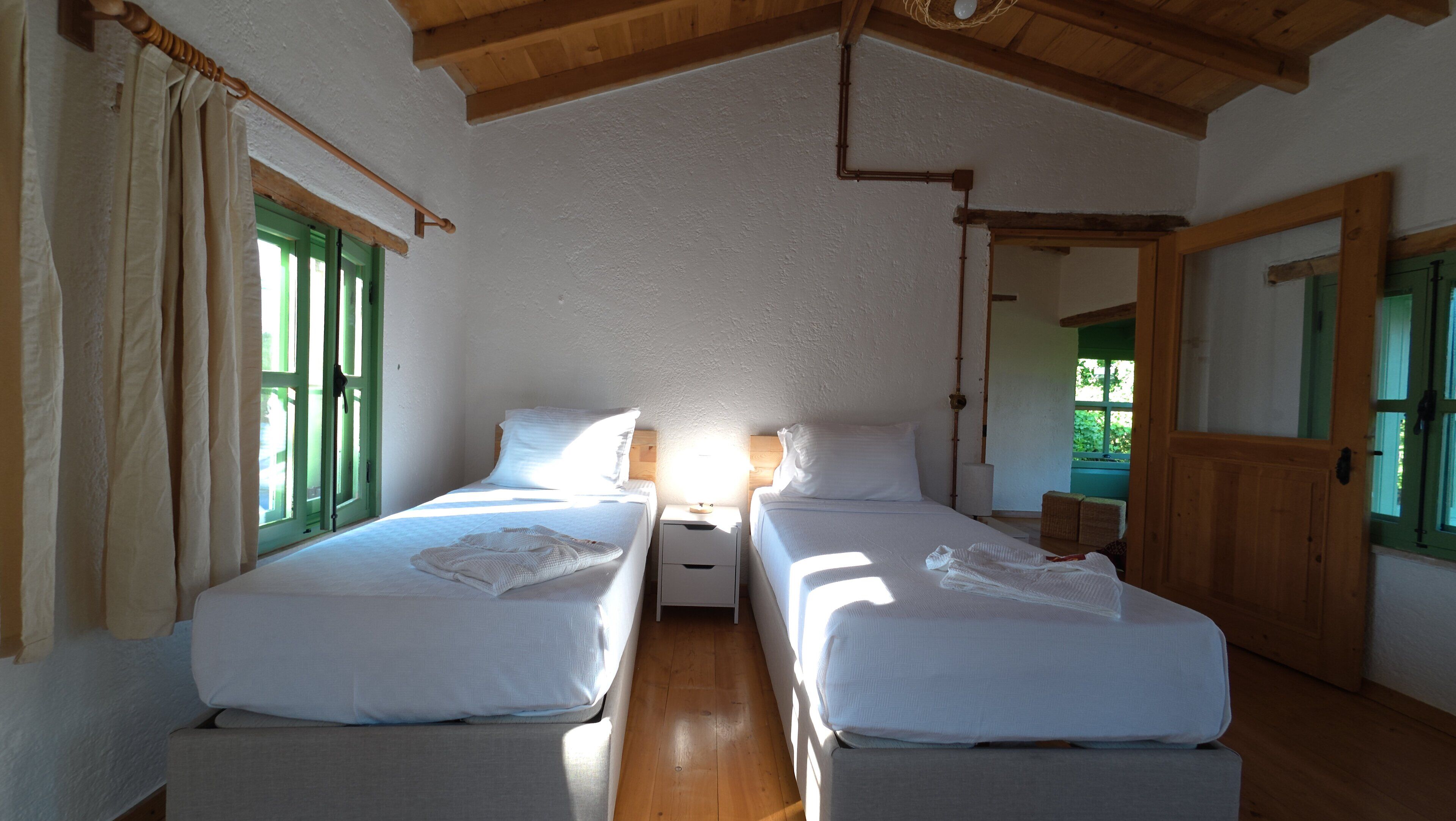 A room with two beds, a wooden ceiling, and a window with green shutters.