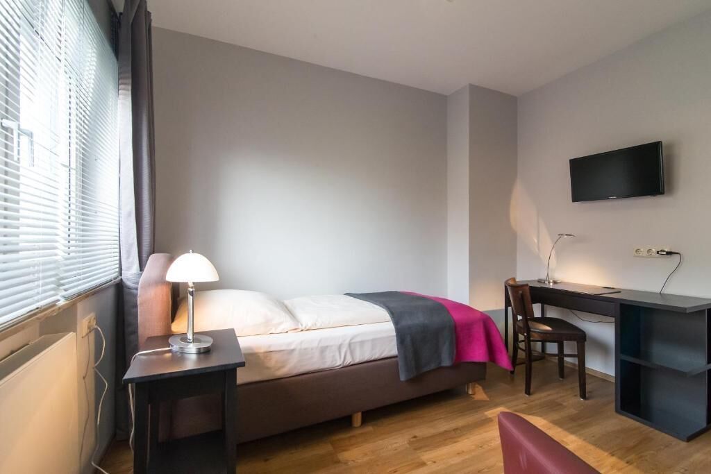 Un dormitorio con cama, mesita de noche, escritorio, silla y televisor en la pared.
