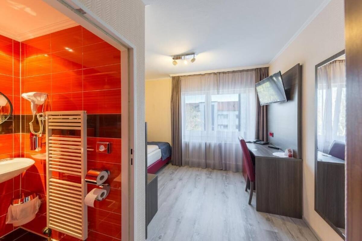 Een moderne hotelkamer met een badkamer met rode tegels, een bed met hoofdeinde, een bureau met stoel en een aan de muur bevestigde televisie.