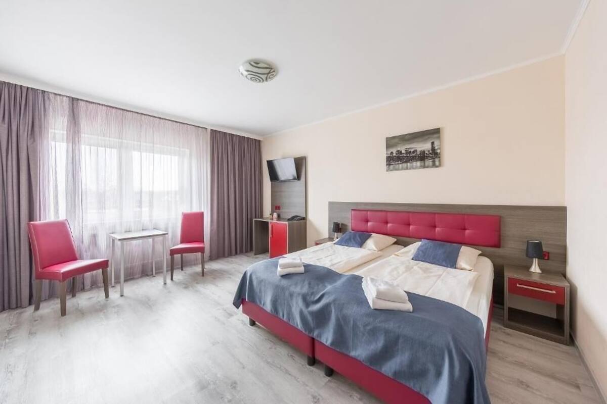 Een hotelkamer met een groot bed, twee rode stoelen, een klein tafeltje, een televisie en een schilderij aan de muur.