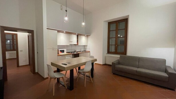 Une cuisine moderne avec des meubles blancs, une table à manger avec des chaises, un canapé gris et des luminaires suspendus.