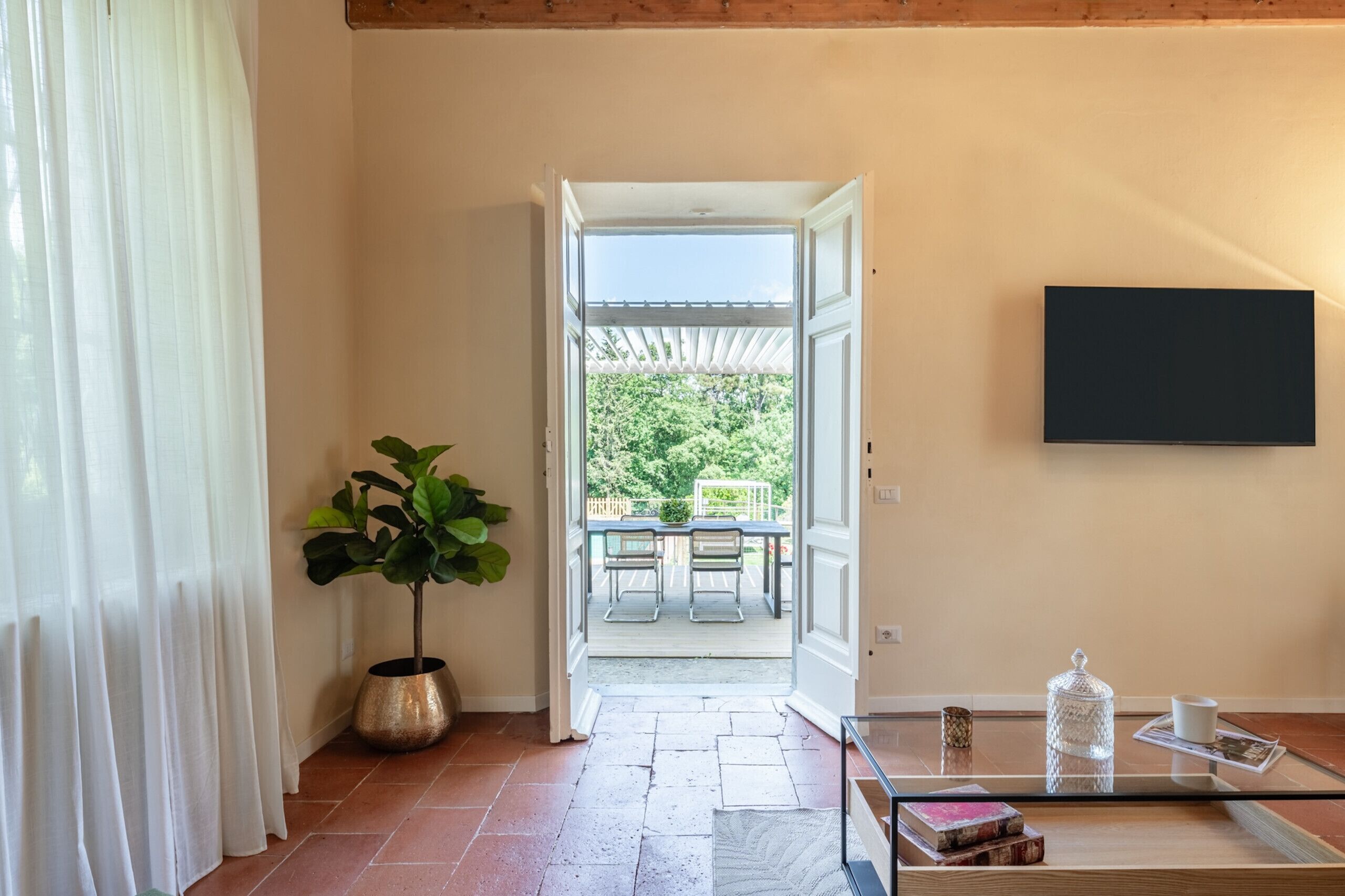 Una habitación con una mesa de centro de vidrio, una planta en maceta, un televisor montado en la pared y vistas a un patio exterior con mesa y sillas.