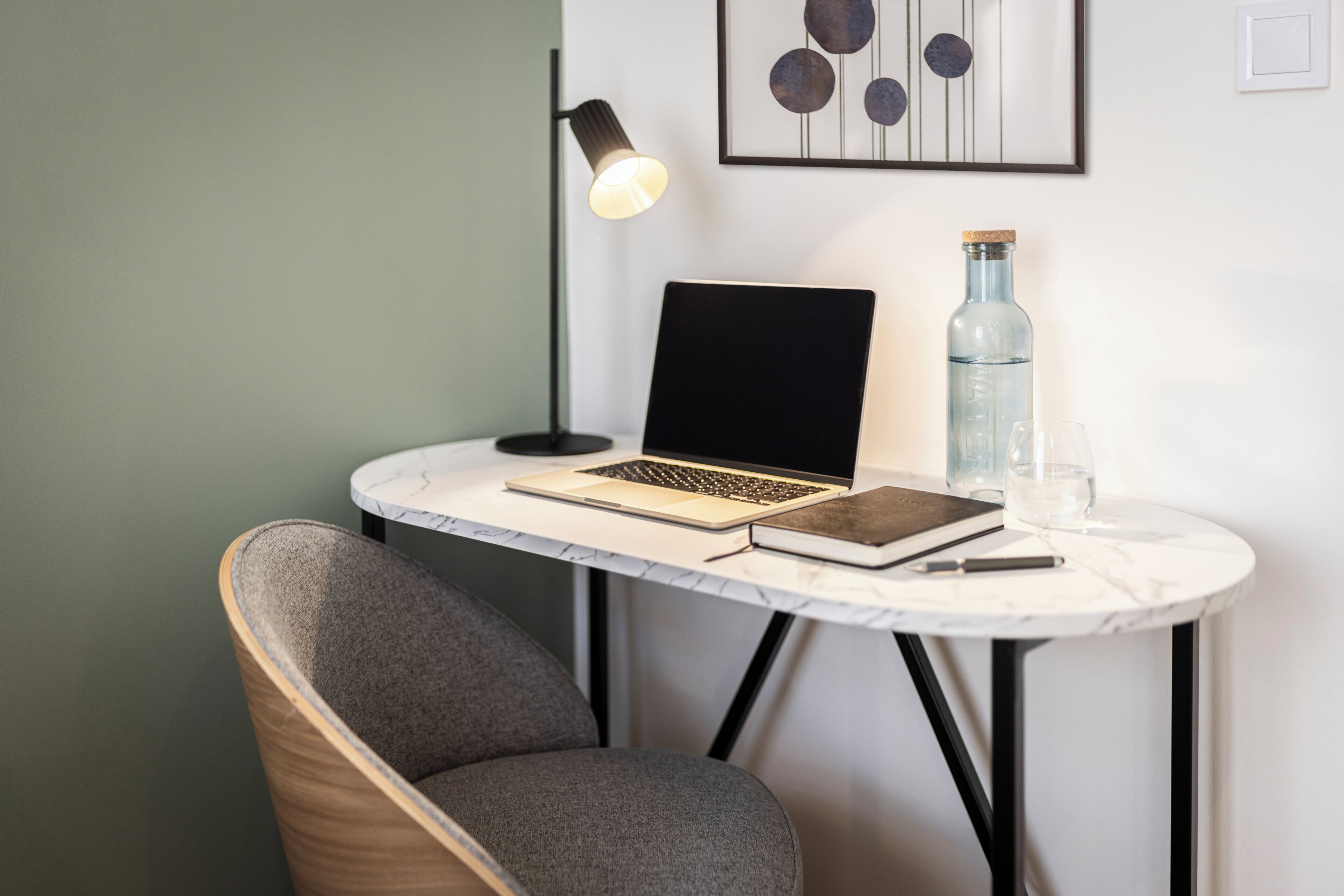 Un espace de travail avec un ordinateur portable, un livre, un verre d’eau et une bouteille sur une petite table ronde.