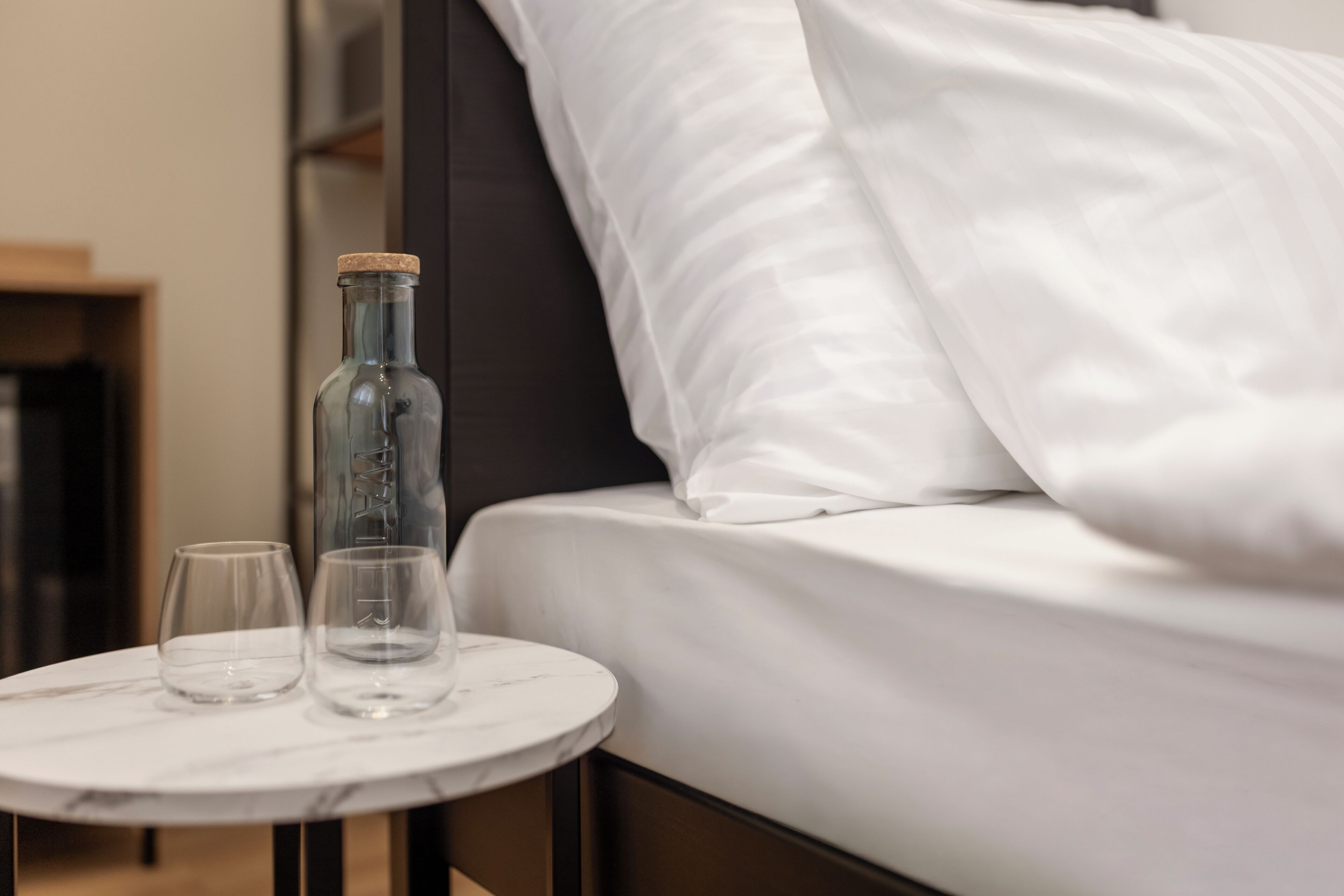 A bed with white bedding, a small round table with a glass bottle and two glasses, and a dark-colored pillow.