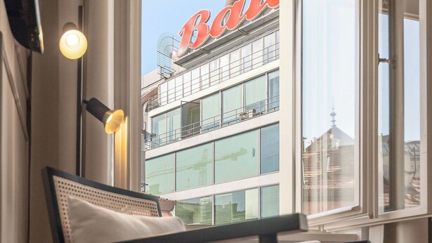 A modern interior with a sofa, a table with a glass, and a view of a building with a red sign. - Youngmann House (Prague)