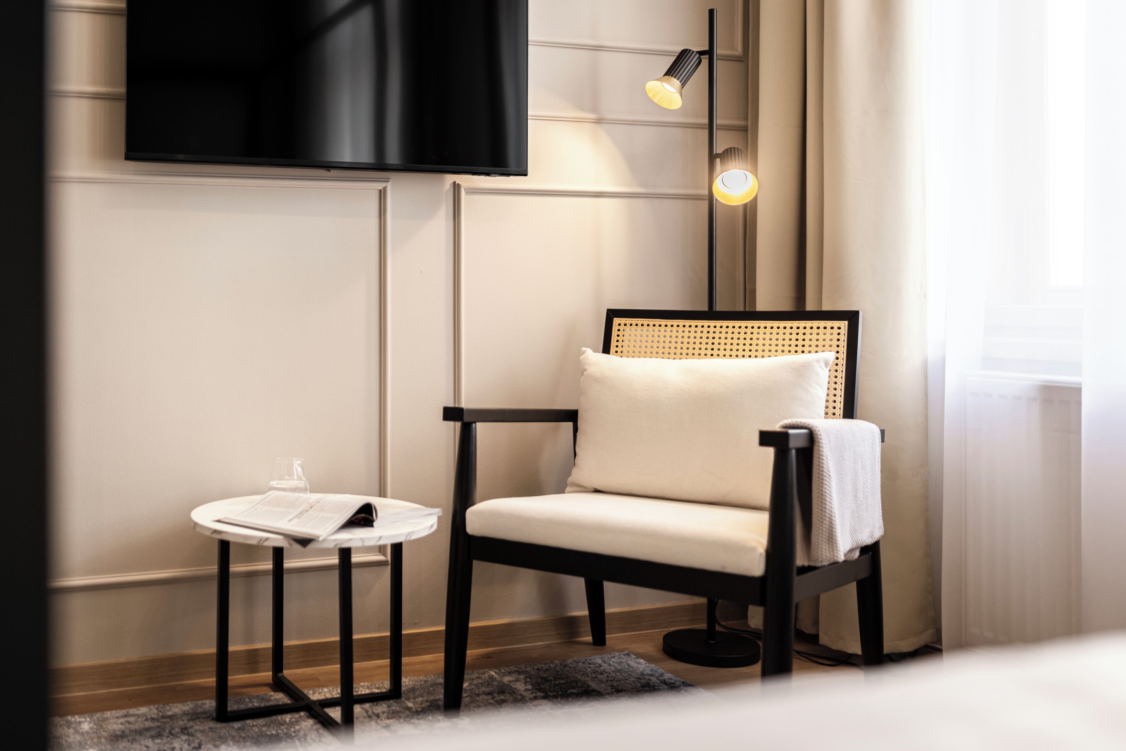 A modern room with a flat-screen TV mounted on the wall, a single chair with a woven backrest, a small round table with a menu, and a floor lamp.