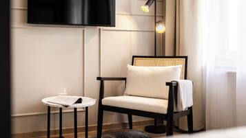A modern room with a flat-screen TV mounted on the wall, a single chair with a woven backrest, a small round table with a menu, and a floor lamp.