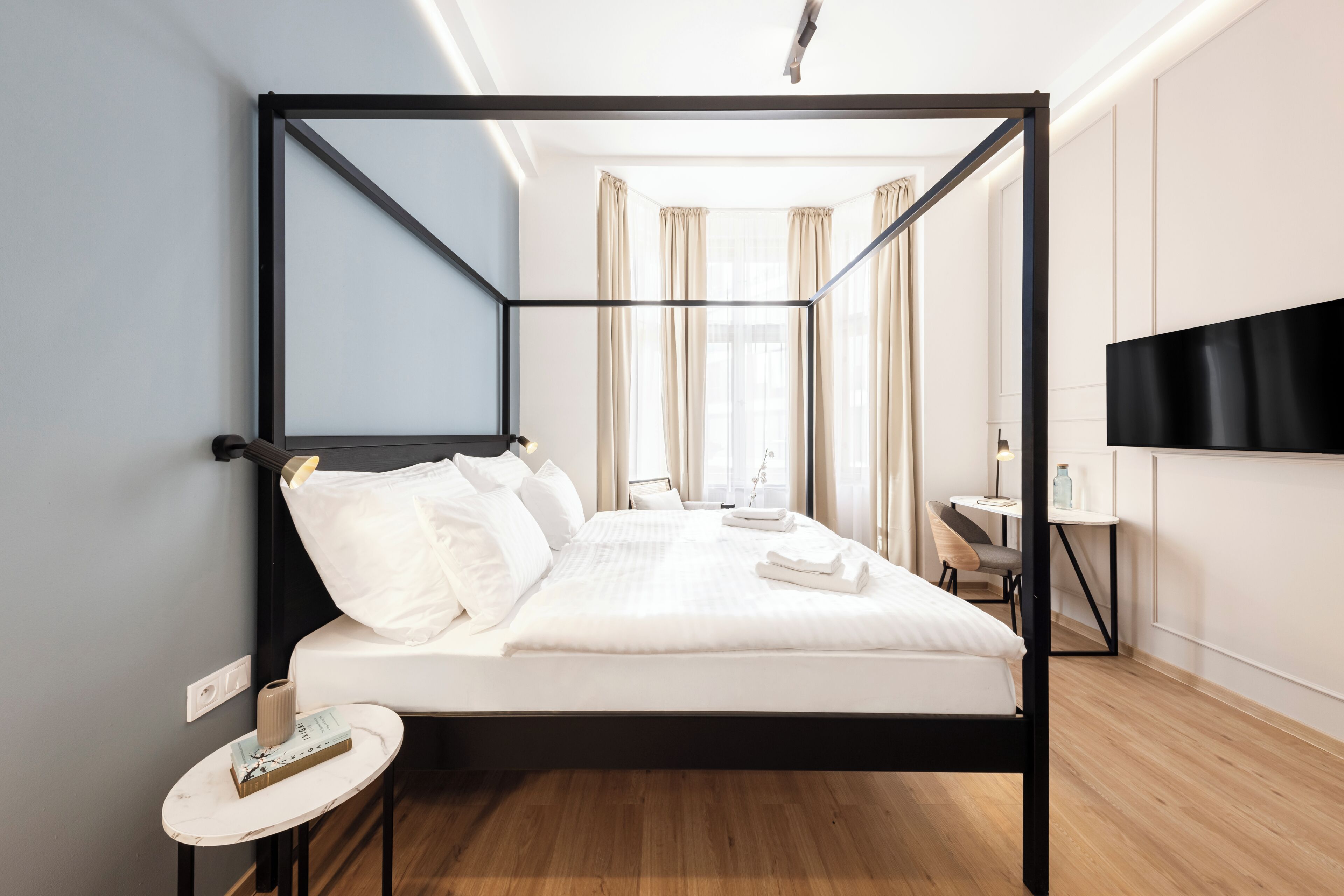A modern hotel room with a four-poster bed, a small round table, a TV mounted on the wall, and a view of the room's interior.
