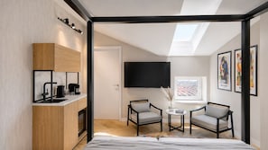 A modern room with a bed, a kitchenette, a TV, two chairs, a small table, and a window with a city view. - Youngmann House (Prague)