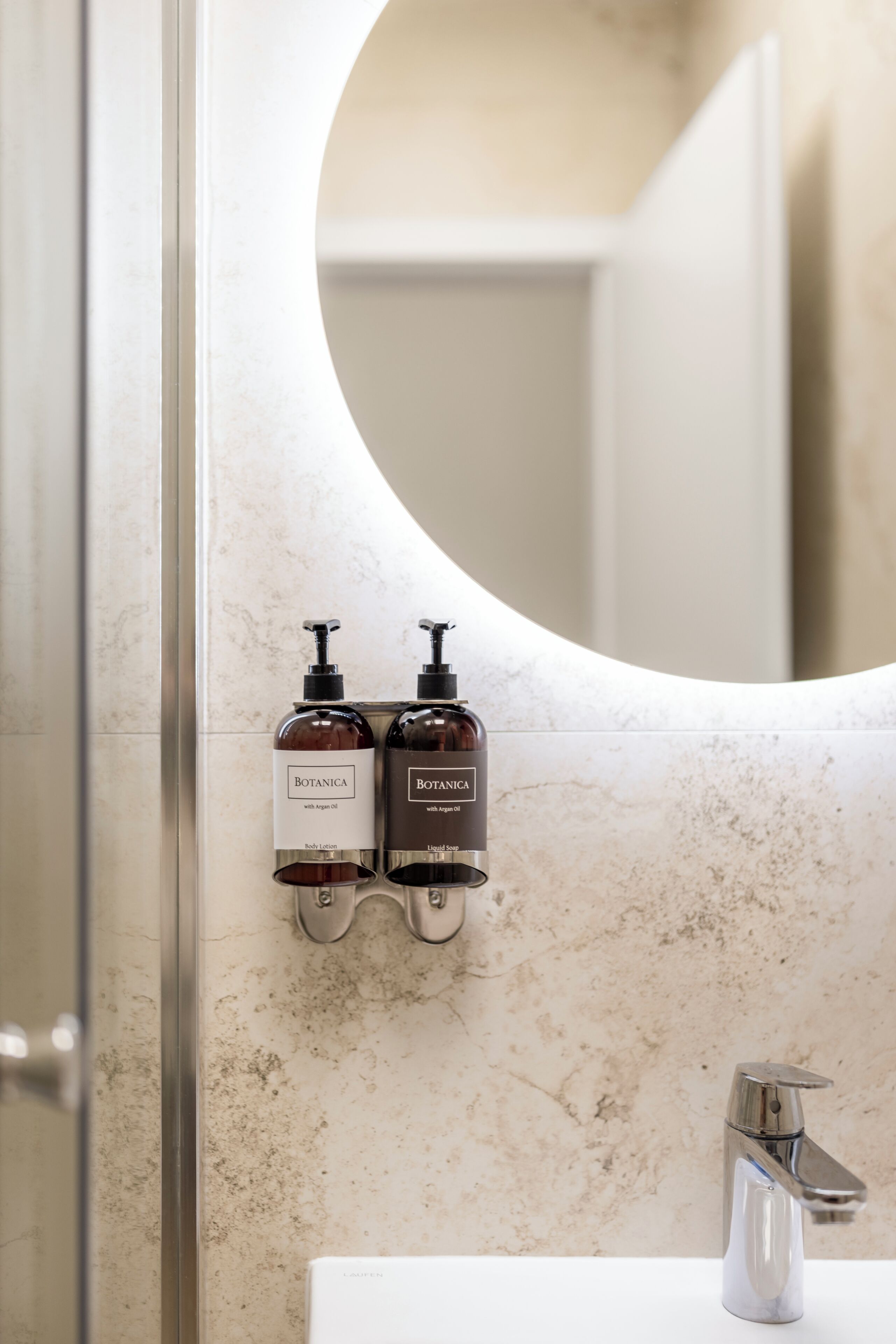 A bathroom with a marble wall, a round mirror, and two bottles of body lotion and liquid soap.