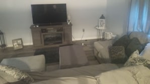 A living room with a flat-screen TV on a stand, a fireplace, a side table with a lantern, and a sofa with cushions. - Nice 1-bedroom cottage with AC in charming West Liberty (West Liberty)