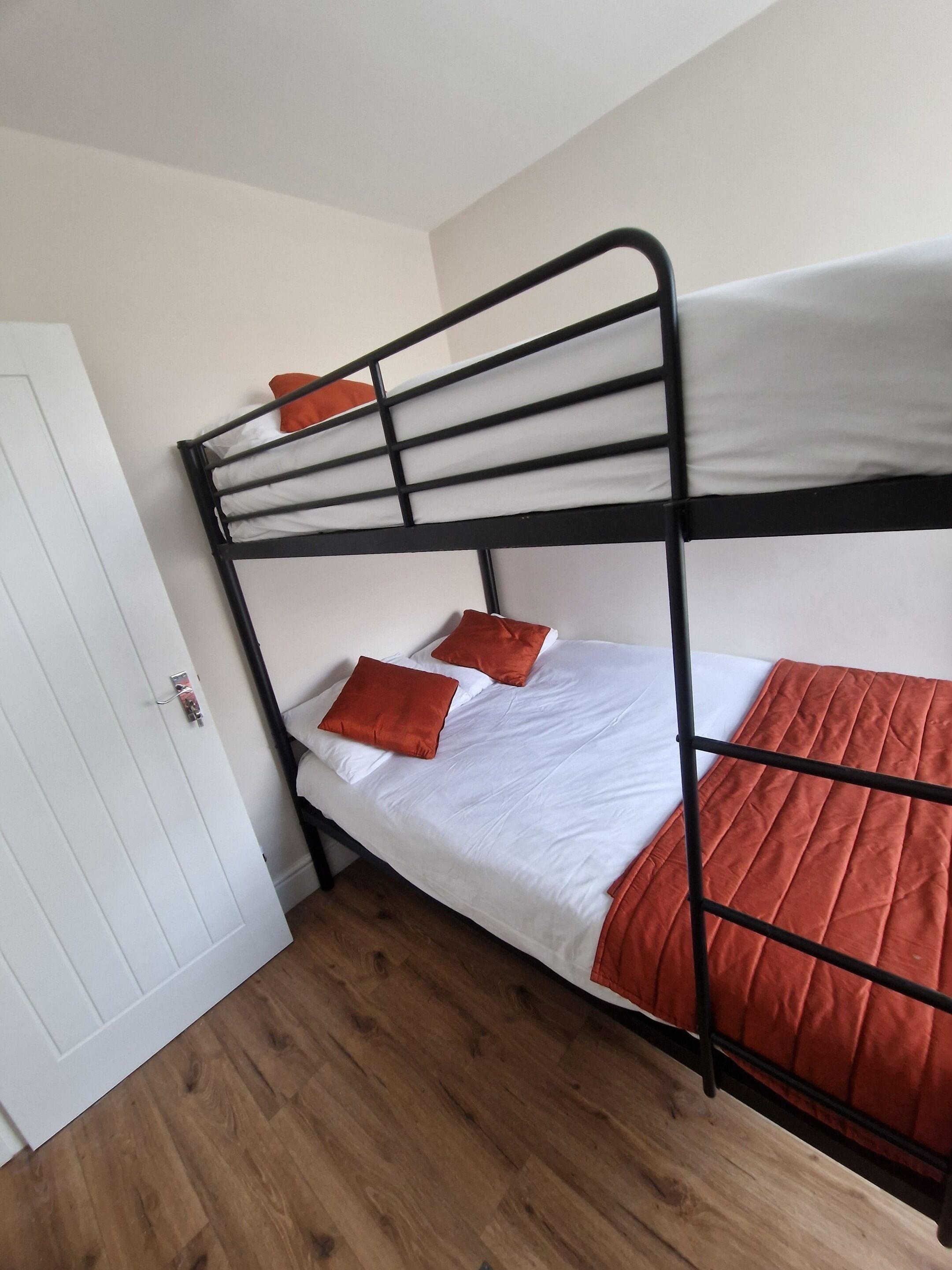 A bunk bed with white bedding and orange pillows, a wooden floor, and a white door.