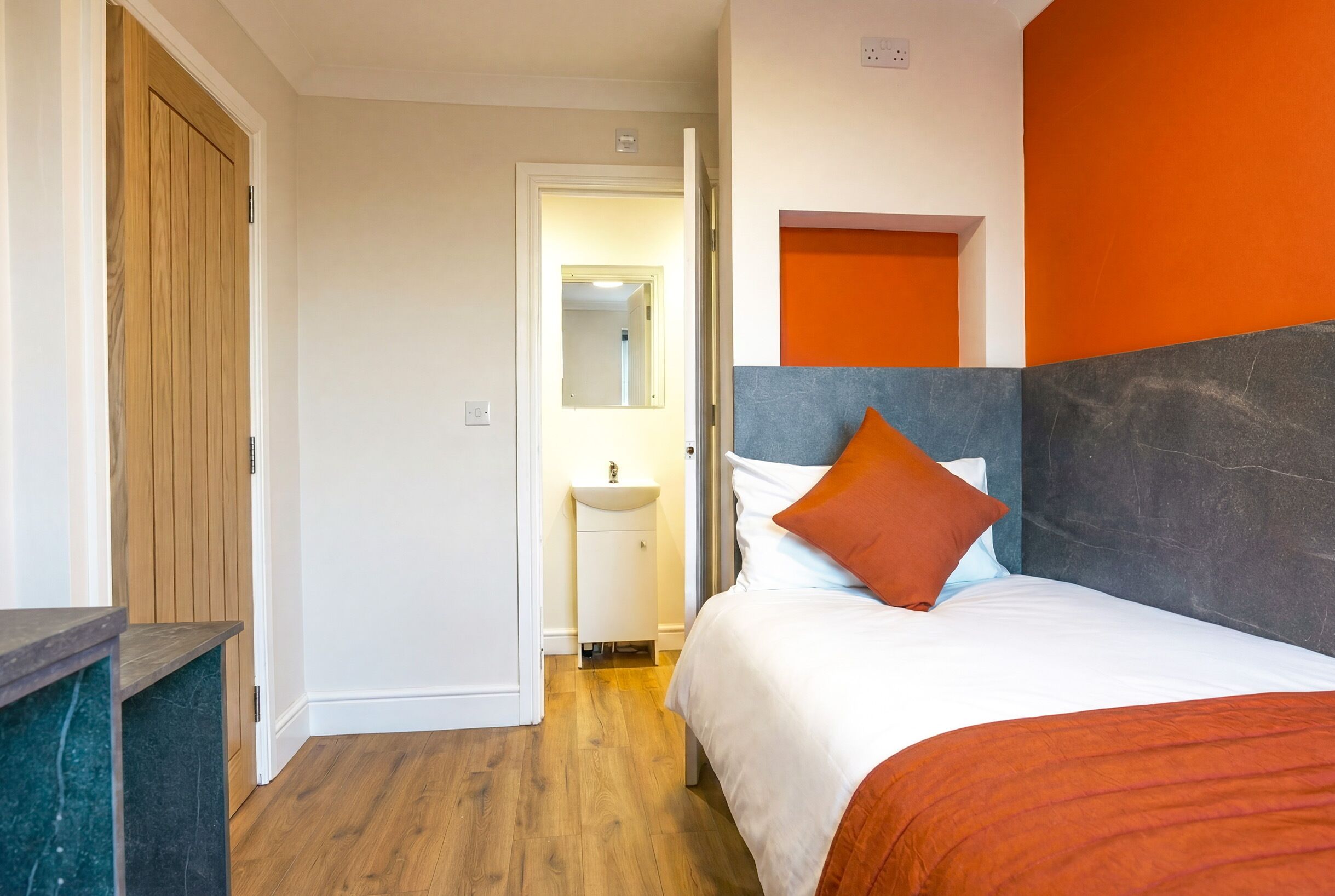 Habitación de hotel con puerta de madera, cama con ropa de cama blanca y naranja, un lavamanos pequeño y un espejo.