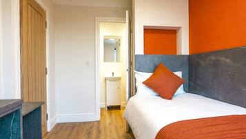 Habitación de hotel con puerta de madera, cama con ropa de cama blanca y naranja, un lavamanos pequeño y un espejo.