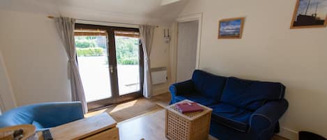 A living room with a blue sofa, wooden coffee table, and a wooden chair. There is a door leading outside and a window with curtains.