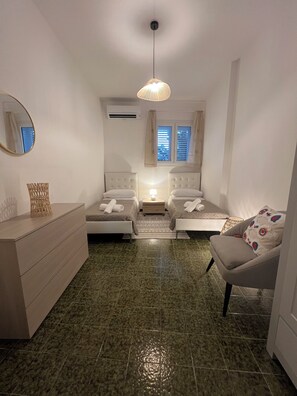 Een slaapkamer met twee bedden, een kaptafel, een stoel, een hanglamp en een raam met jaloezieën.