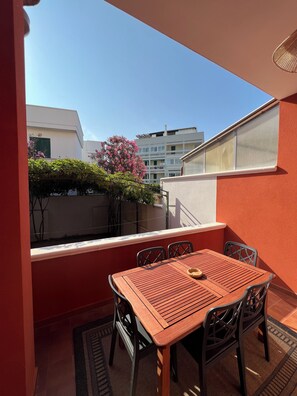 Een balkon met een houten tafel en stoelen, uitzicht op gebouwen en een bloeiende roze plant.