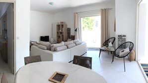 A modern living room with a sofa, a dining table, and a kitchen area visible through an open door. - Appartement proche du centre historique, terrasse, clim et garage privé fermé. (Arles)