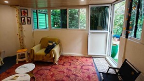 Ein Zimmer mit Holzbalkendecke, einem roten Teppich mit Muster, einem gelben Sofa, einem kleinen Hocker, einem weißen Tisch und einem schwarzen Stuhl.