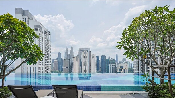 Une piscine à débordement située sur le toit, avec des chaises longues, entourée de verdure et offrant une vue panoramique sur les gratte-ciel d’une ville moderne.