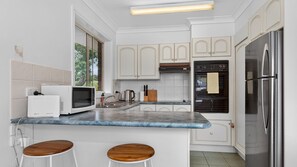 A modern kitchen with white cabinets, a built-in oven, a microwave, and a stainless steel refrigerator. There are two wooden stools at the counter. - Coastal Comfort at Malua Bay (Malua Bay)