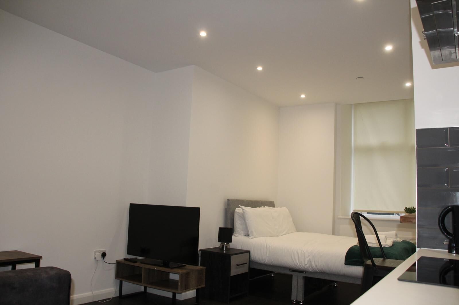 A modern hotel room with a bed, a TV on a wooden stand, a bedside table, and a window with a view.