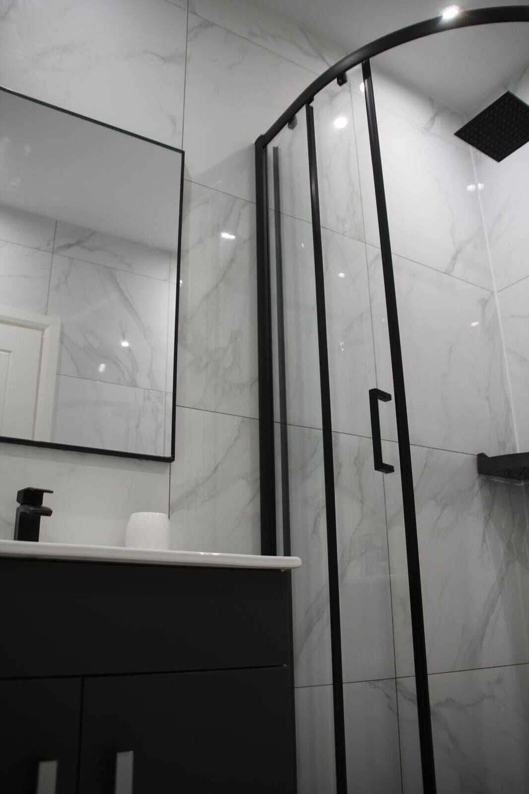 Un baño moderno con cabina de ducha de vidrio curvo, pared revestida de mármol y tocador blanco con grifo negro.