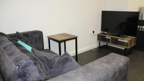 Ein Wohnzimmer mit grauem Sofa, einem kleinen Holztischchen, einem Fernseher-Schrank und einem Flachbildfernseher.