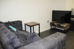 Ein Wohnzimmer mit grauem Sofa, einem kleinen Holztischchen, einem Fernseher-Schrank und einem Flachbildfernseher.