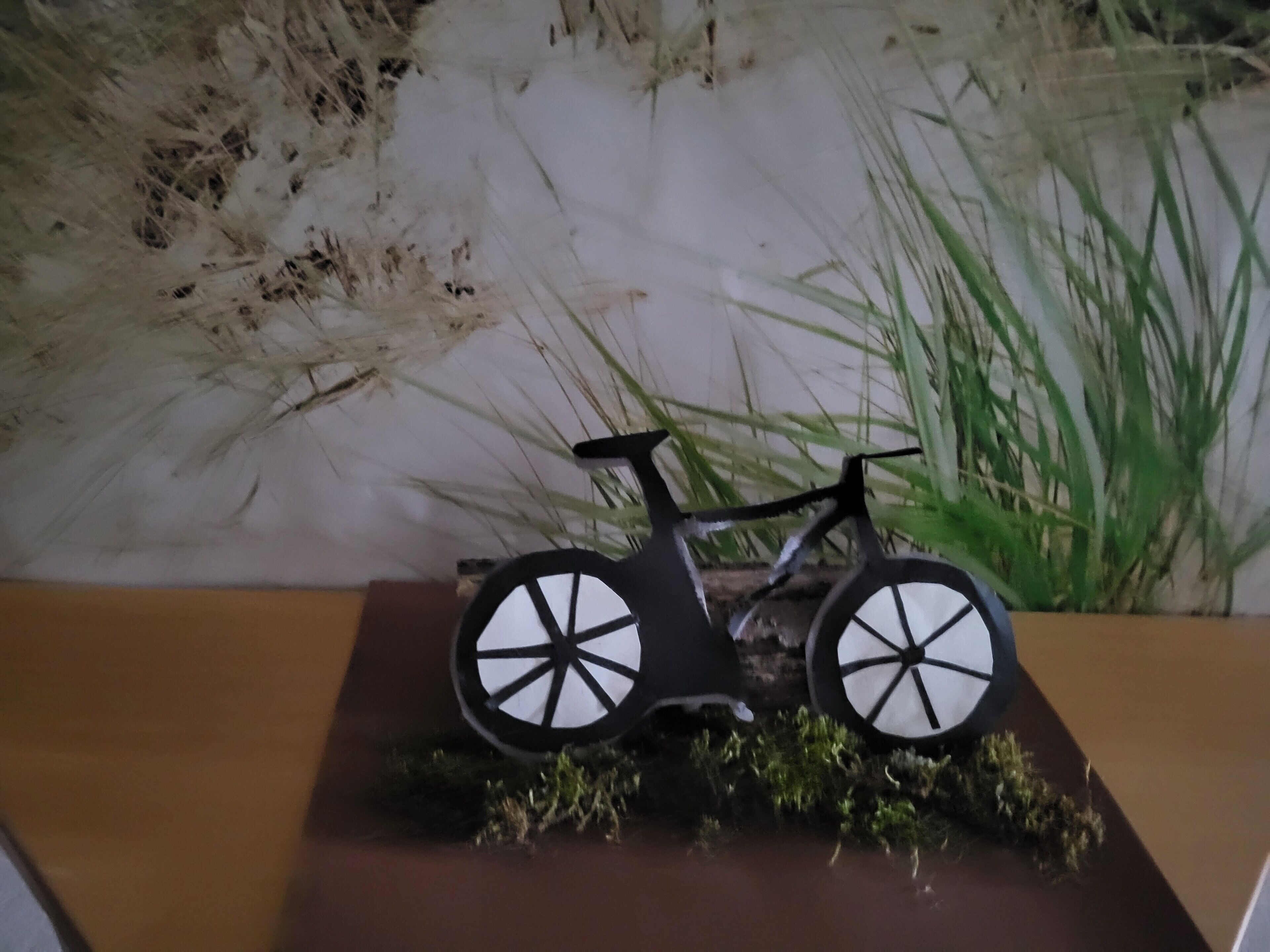 Ein Modellfahrrad auf einem Holzfuß mit Gras und einer Wand im Hintergrund.