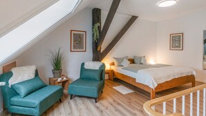 Une chambre avec un plafond en pente, un lit en bois, deux fauteuils turquoise, une petite table d’appoint et une plante en pot.