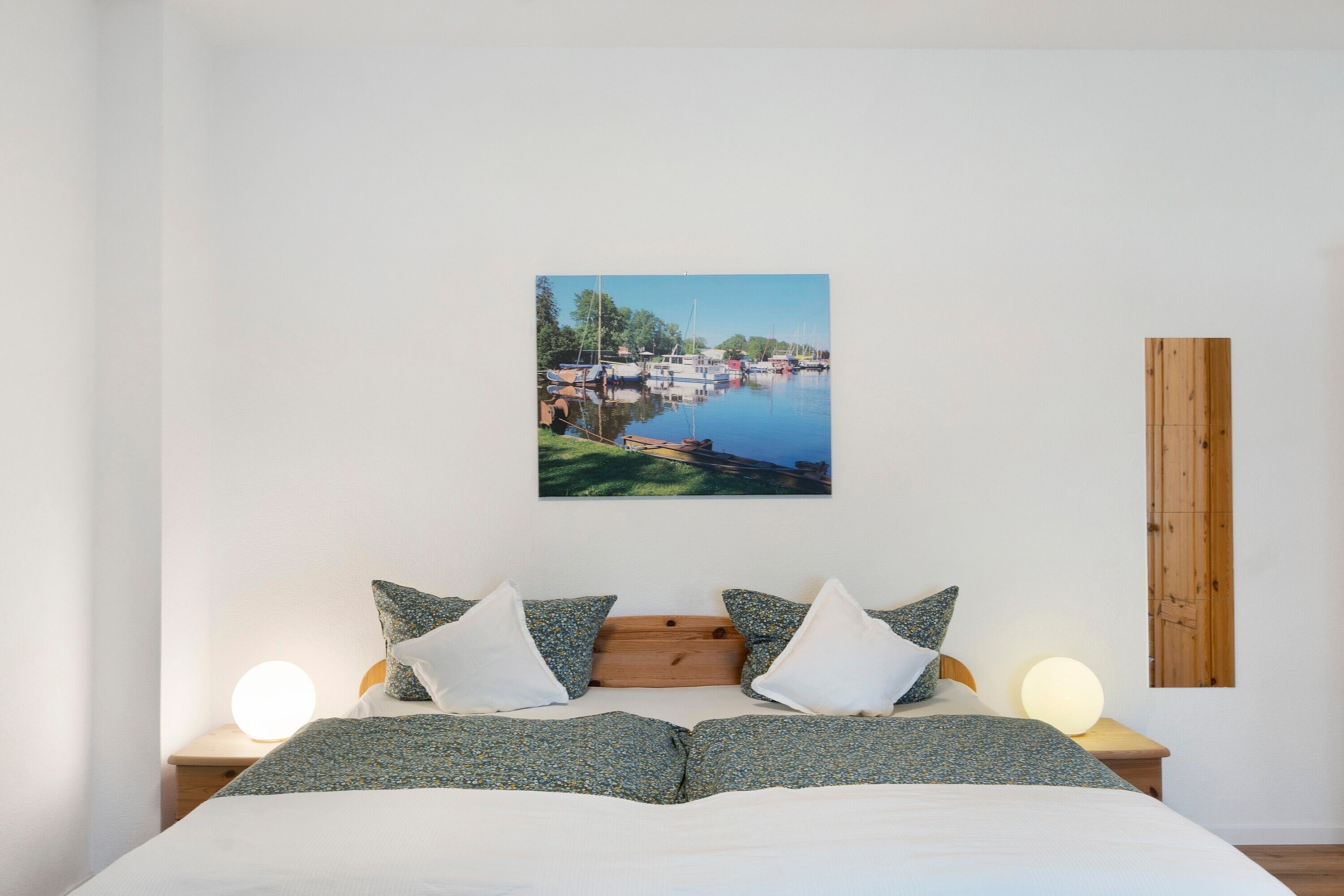 Ein Bett mit Holz-Kopfteil, zwei Nachttische mit Lampen und ein Wandbild mit Seeblick.