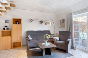 Ein modernes Wohnzimmer mit Sofa, Sesseln, Couchtisch, Bücherregal und einem großen Fenster.