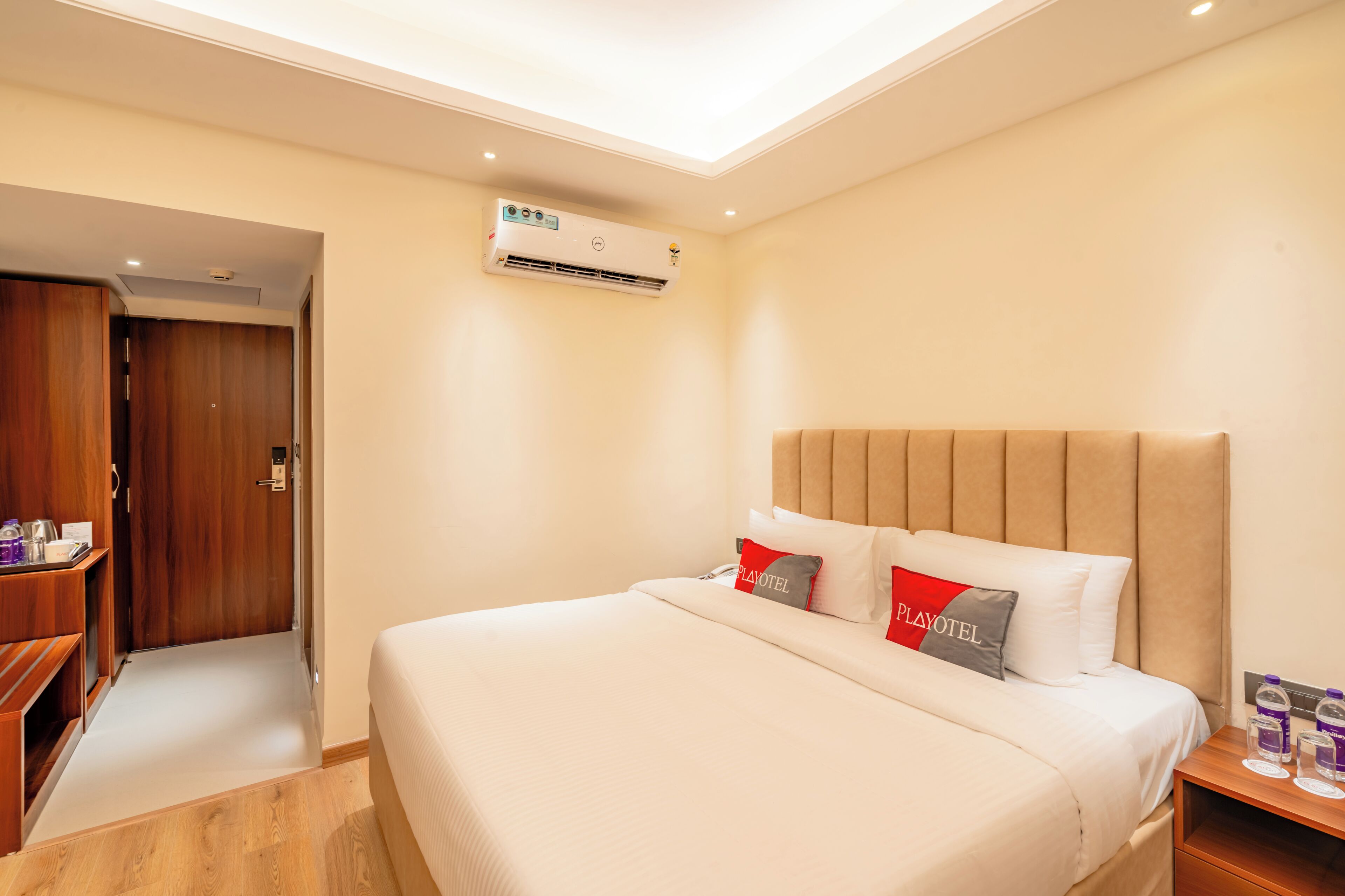 A hotel room with a bed, a wooden wardrobe, an air conditioning unit, and a nightstand with a water bottle and glasses.