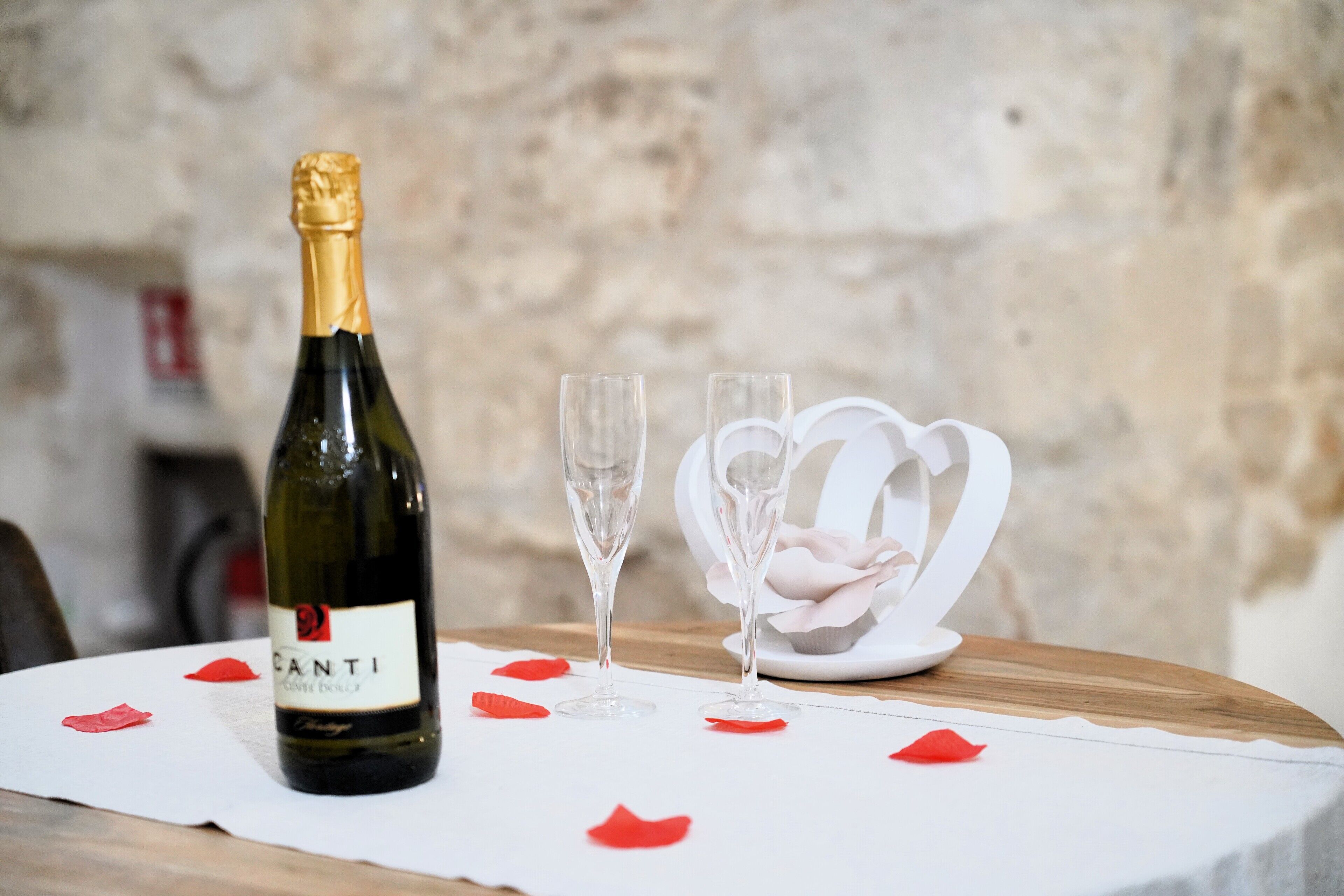 Una bottiglia di vino Canti, due bicchieri e una decorazione a forma di cuore bianco su un tavolo con tovaglia bianca e petali di rosa rossa.