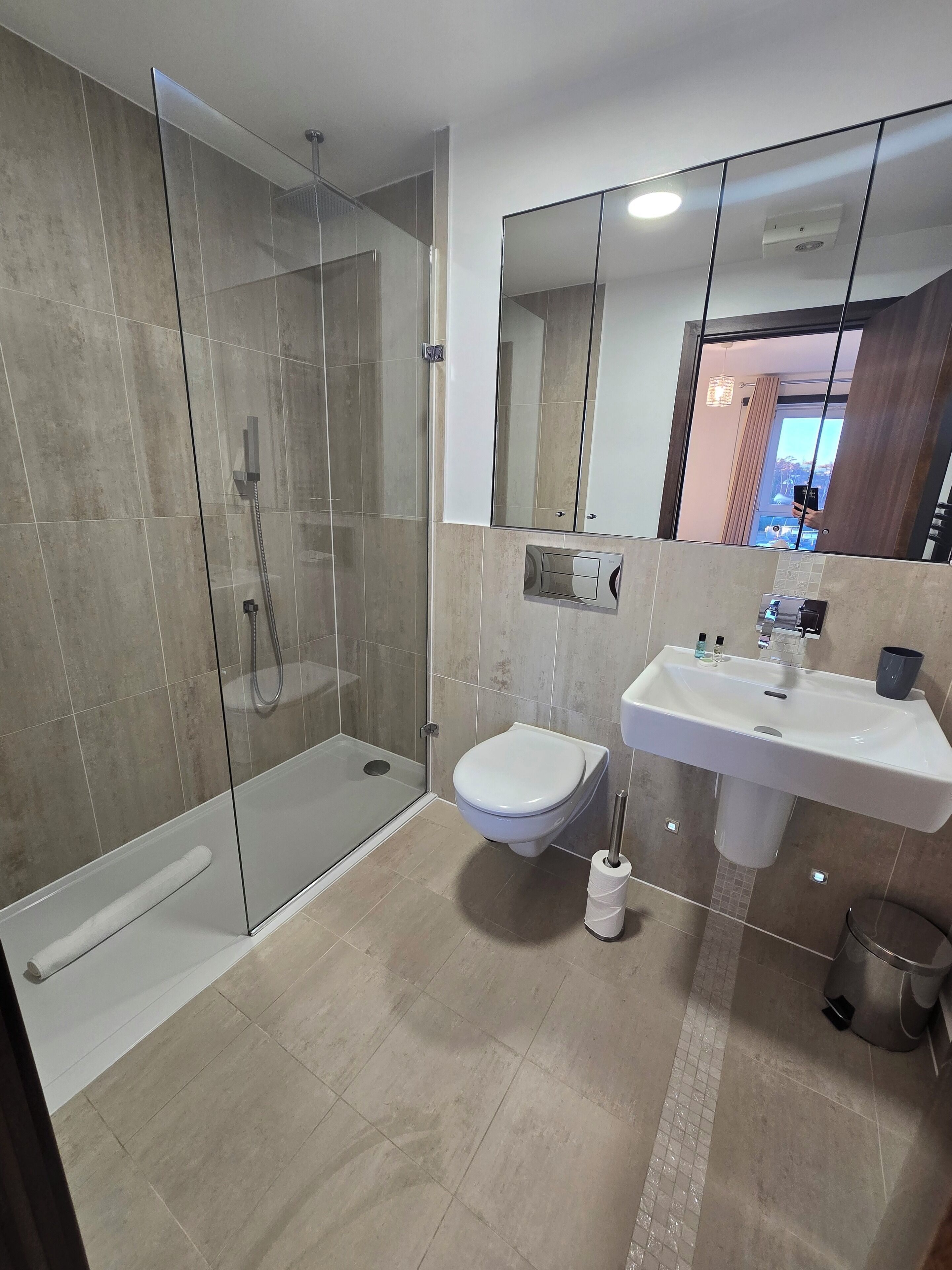 A modern bathroom with a glass-enclosed shower, a white toilet, a sink with a mirror above it, and a trash bin.